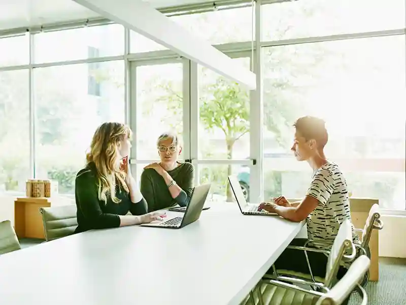 A mature businesswoman leads a discussion in a bright, windowed office conference room.